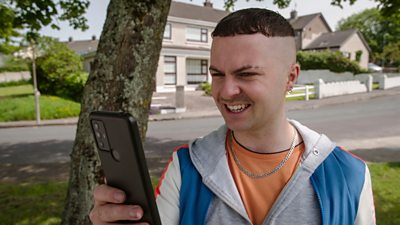 Conor MacSweeney (Alex Murphy) smiles as he looks at a smartphone.