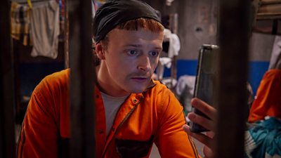 Photo of Jock (Chris Walley) looking at a smartphone as he sits in a prison cell wearing orange overalls. 
