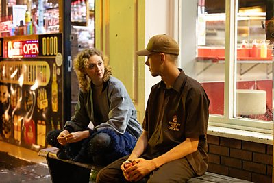 A young man talks to a young woman on a bench in a nighttime street