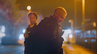 A male police officer in a night time street. A female police officer behind him looks concerned