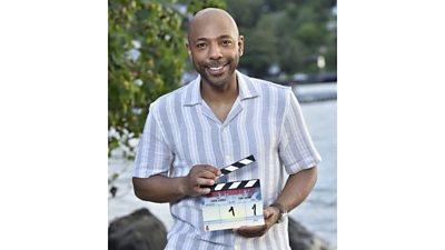 Don Gilet smiles to camera and holds a clapperboard for a new series of Death in Paradise 
