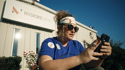 woman with a headband and sunglasses on looking at her phone. cabin in background with sign mvskoke media on it