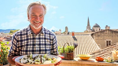 Marcus Wareing smiling and holding a plate of food with rooftops and blue sky in background