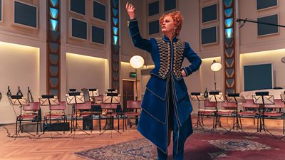 Jinkx Monsoon as Maestro in The Devil's Chord. Maestro stands in the middle of a recording studio, surrounded by empty orchestra seats, poised to strike.
