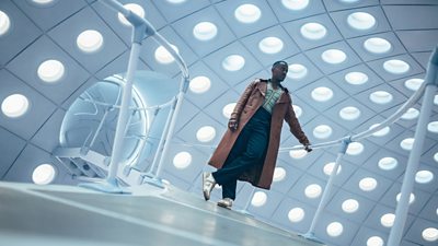 Ncuti Gatwa as The Doctor, standing in the TARDIS, captured from below. The Doctor wears a long brown coat, dark trousers, trainers and a knitted vest.