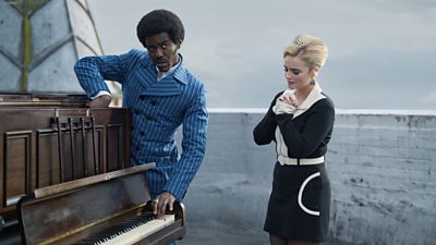 The Doctor and Ruby Sunday stand by a piano. The Doctor plays some keys as Ruby watches on.