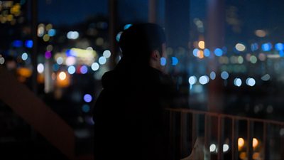 A sillouette of a person stood outside at nighttime, a city skyline is blurred in the background.