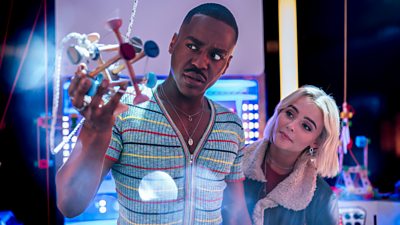 Photo of Ncuti Gatwa and Millie Gibson as the Doctor and Ruby Sunday. The Doctor looks at a hanging model with concern, as Ruby looks over his shoulder.
