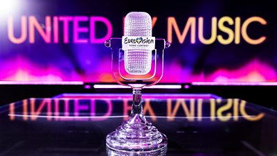 The Eurovision 2024 trophy, a crystal microphone, sits on the stage. United By Music is written in bright neon colours on the screen behind the stage.