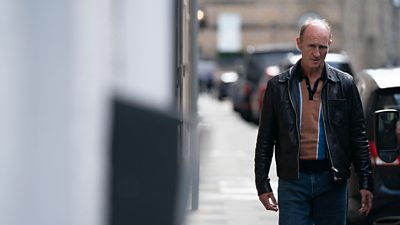 Stuart Bowman (Ger Cafferty) walks along a street and looks towards camera. 