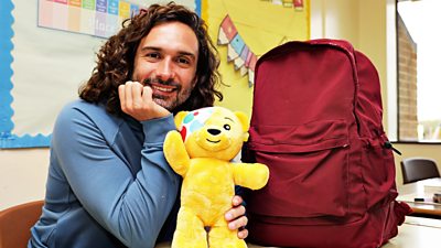 Joe Wicks smiles to camera holding a Pudsey soft toy, sat beside a red backpack. 