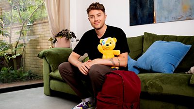 Roman Kemp smiles to camera holding a Pudsey soft toy with a  red backpack at his feet. 