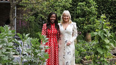 Angellica Bell and Nicki Chapman stand in a garden full of plants, flowers, and trees and smile to camera. 