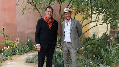 Monty Don and Joe Swift stand in a small, green space surrounded by trees and flowers.