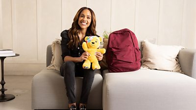Alex Scott smiles to camera holding a Pudsey soft toy, sat beside a red backpack. 
