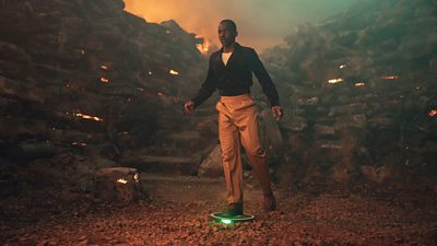 Ncuti Gatwa's Fifteenth Doctor stands on an illuminated land mine, looking shocked. The Doctor balances on one leg.
