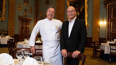 John Torode and Gregg Wallace smile to camera from a large banquet hall filled with dining tables. 