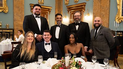 Poppy O'Toole, Big Has, Stu Deeley, Aktar Islam, Keziah Whittaker, Jonny Lake, Nathan Davies smile to camera from a table at the MasterChef 20th Anniversary Dinner.