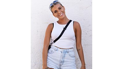 hannah wearing jean shorts and a white top smiling with head slightly tilted and sunglasses on her head