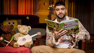 Photo of Zayn Malik holding a book in the CBeebies Bedtime Story studio, next to two teddy bears with pictures of musical instruments. 