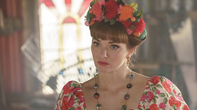 Gabby (Eleanor Tomlinson) looks at something off-camera wearing a headband with large, 3-D flowers