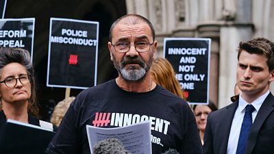 Andy Malkinson outside the Court of Appeal in front of microphones. A small crowd behind him hold signs reading: "police misconduct" and "innocent and not the only one"