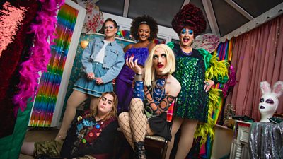 group of people in a colourful room. three in drag and two wearing colourful outfits. rainbow draping and feather boas in the background