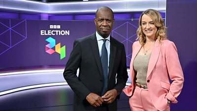 Clive Myrie and Laura Kuenssberg stand in a studio with Election 2024 on a screen behind them. Clive wears a dark suit, shirt and tie, while Laura wears a lighter pink suit with a gray top.