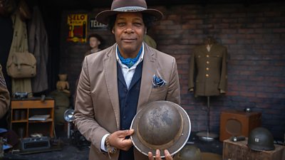 Bargain Hunt presenter Danny Sebastian stands holding a metal helmet with a wide brim, surrounded by war memorabilia including coats, bags, uniforms, old flashlights and an old radio