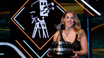 mary earps smiling holding the sports personality of the year trophy. background of neon orange and blue lights with a camera 