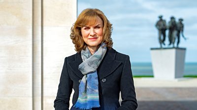 Fiona Bruce stands outdoors wearing a dark coat and blue patterned scarf. A statue of three soldiers is slightly blurred in the background.