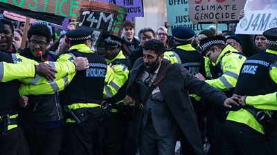 Adeel Akhtar pushes through a line of police officers. The officers hold back a large group of protestors with signs with text reading: “Climate genocide”