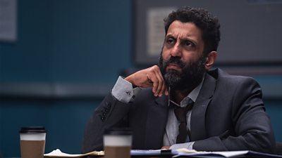 Adeel Akhtar looks at someone off-camera with a concerned expression. Files and coffee cups sit on the table in front of him. 