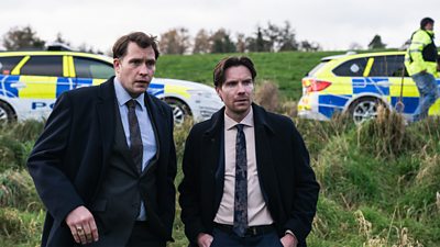 Tom Padley and Joe Dempsie stand in a field wearing suits. Police cars are visible in the background. 