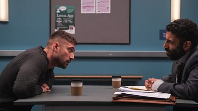 Michael Socha leans across the table towards Adeel Akhtar in a police interview room. 