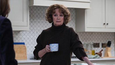 Francesca Annis stands in a kitchen holding a coffee cup. She looks to someone off-screen. 
