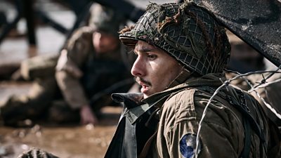 A soldier on the battlefield, sitting down, looking shocked. He wears a helmet and uniform. In the background, another soldier crouches on the ground.