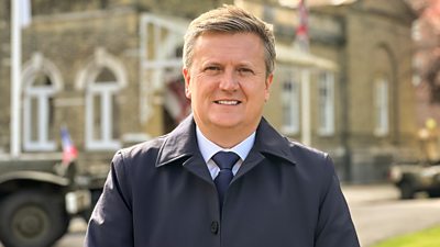 Aled Jones stands outdoors wearing a white shirt and dark blue tie, with a dark navy coat.