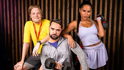 two men and one woman looking to camera. One in front holding a dumbbell and wearing a. zip up hoodie. woman to his right wearing shorts and purple top holding a kettlebell up to her shoulder and leaniing on man. Man to the left leaning on man with other arm resting on his leg