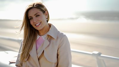 Amy Dowden stands by a coastline, wearing a light jacket over a pink jumper with a lace collar.