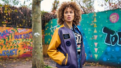 Mia (Michelle De Swarte) stands in front of two walls covered in colourful graffiti. 