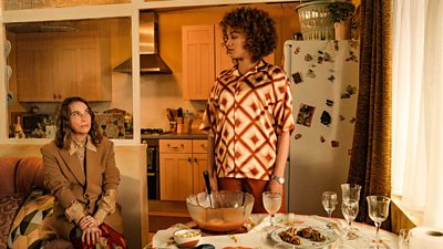 Jemima (Eva Pope) sits on the edge of a sofa and looks up at Mia (Michelle De Swarte). The pair are in an open-plan kitchen, beside a dining table covered in food and empty glasses. 