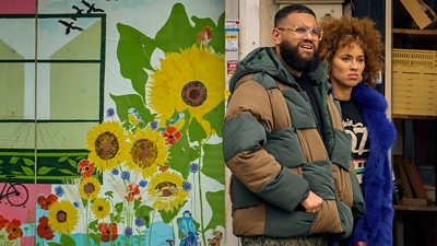 Marc (Jamal Maddox) stands beside Mia (Michelle De Swarte) in front of a wall covered in a colourful mural. 