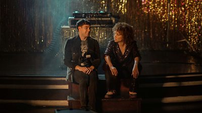 David (Joseph Ollman) and Mia (Michelle De Swarte) sit on the edge of a stage. A DJ turntable sits on the stage behind them, in front of a glittery backdrop. 