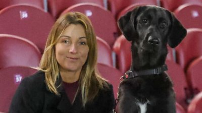 A black dog sits next to a blonde woman at a football stadium with red seats.
