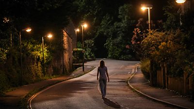 India Lille Davies as Andie Bell walks along a road alone at night. The streetlights illuminate the scene. 