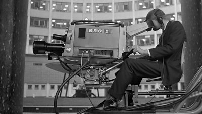 A cameraman seated operating a large broadcast camera with ̳ 2 on the site. The doughnut at Television Centre is behind him