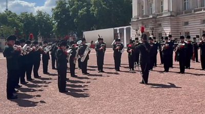 Two semi-circle lines of guards play instruments outside Buckingham Palace with sun shining down on them