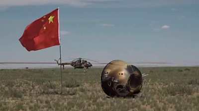 Lunar probe in a field, next to Chinese flag and with helicopter in bacground