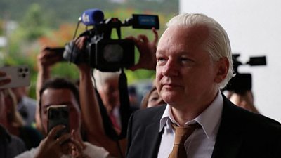 Julian Assange dressed in a suit and tie surrounded by cameras